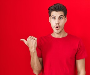 Young hispanic man standing over red background surprised pointing with hand finger to the side,...
