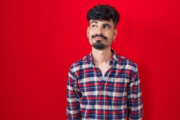 Obraz premium Young hispanic man with beard standing over red background smiling looking to the side and staring away thinking.