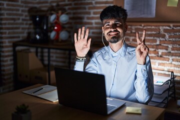 Young hispanic man with beard working at the office at night showing and pointing up with fingers number seven while smiling confident and happy.