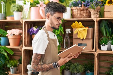 Young hispanic man florist reading notebook with relaxed expression at flower shop - obrazy, fototapety, plakaty