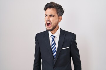 Young hispanic man with tattoos wearing business suit and tie angry and mad screaming frustrated and furious, shouting with anger. rage and aggressive concept.