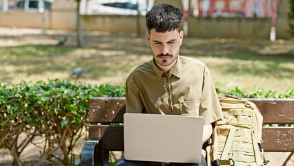 Young hispanic man tourist using laptop sitting on bench at park