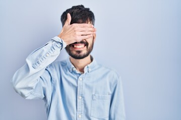 Young hispanic man with beard standing over blue background smiling and laughing with hand on face...