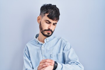 Young hispanic man with beard standing over blue background checking the time on wrist watch,...