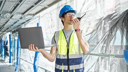 Young hispanic man architect talking on walkie-talkie using laptop at construction place