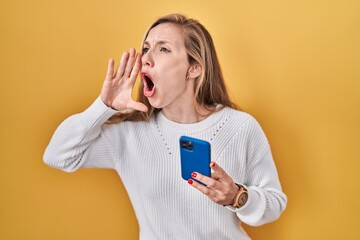 Young blonde woman using smartphone typing message shouting and screaming loud to side with hand on mouth. communication concept.