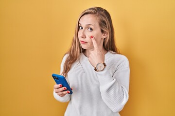 Young blonde woman using smartphone typing message pointing to the eye watching you gesture, suspicious expression - obrazy, fototapety, plakaty