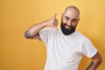 Young hispanic man with beard and tattoos standing over yellow background smiling doing phone gesture with hand and fingers like talking on the telephone. communicating concepts.