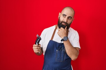 Young hispanic man with beard and tattoos wearing barber apron holding razor with hand on chin thinking about question, pensive expression. smiling with thoughtful face. doubt concept.