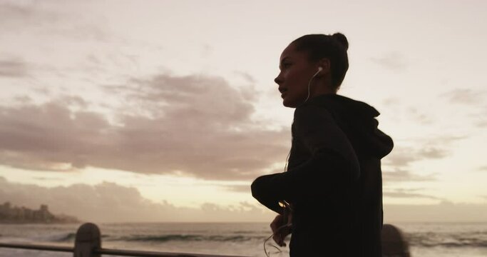 Beach, fitness and woman with phone, earphones or music choice at sea for cardio run on sky background. Smartphone, radio and runner at the ocean with app, playlist or podcast for training motivation