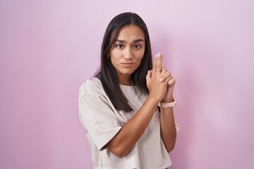 Young hispanic woman standing over pink background holding symbolic gun with hand gesture, playing killing shooting weapons, angry face