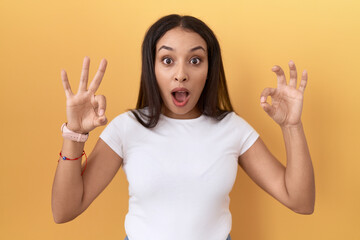 Young arab woman wearing casual white t shirt over yellow background looking surprised and shocked doing ok approval symbol with fingers. crazy expression