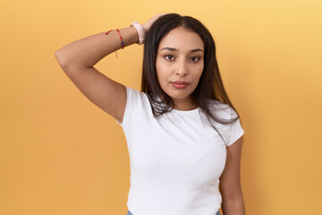 Young arab woman wearing casual white t shirt over yellow background confuse and wondering about question. uncertain with doubt, thinking with hand on head. pensive concept.