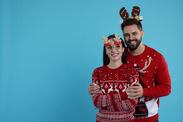 Happy young couple in Christmas sweaters, reindeer headband and funny glasses on light blue background. Space for text