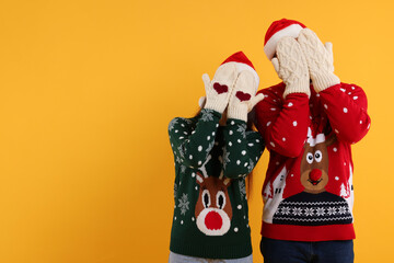 Couple in Christmas sweaters and Santa hats covering faces with hands in knitted mittens on orange background. Space for text