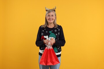 Happy senior woman in Christmas sweater and deer headband with shopping bags on orange background