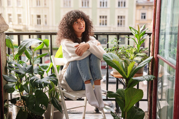 Beautiful young woman relaxing in chair surrounded by green houseplants on balcony