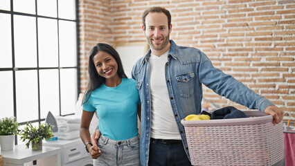 Beautiful couple holding basket with clothes hugging each other smiling at laundry room