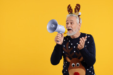 Senior man in Christmas sweater and reindeer headband shouting in megaphone on orange background....
