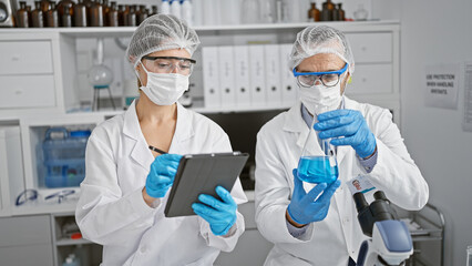 Together in the trenches, two scientists, donned in gloves and masks, scribble on touchpad amidst...