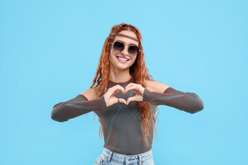 Stylish young hippie woman in sunglasses making heart with hands on light blue background