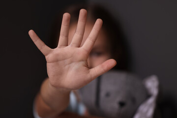 Child abuse. Little girl with toy bunny doing stop gesture on dark background, selective focus - obrazy, fototapety, plakaty