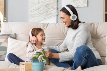 Beautiful mother and her cute little daughter in headphones with mobile phone sitting on sofa at...