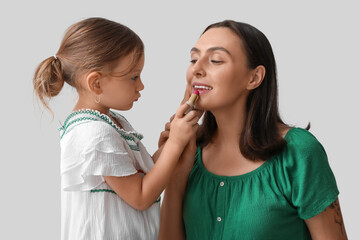 Cute little daughter applying lipstick onto mother's lips on grey background