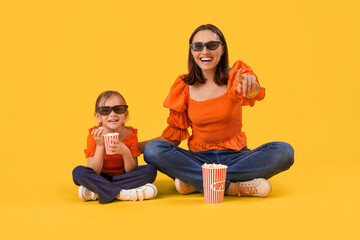 Beautiful mother and her cute little daughter in 3D glasses with buckets of popcorn on yellow background