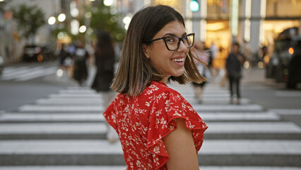 Beautiful hispanic woman in glasses, smiling and looking back at camera, walking across tokyo street crosswalk