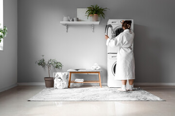 Young woman in white bathrobe putting clothes into washing machine at laundry room