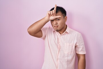 Chinese young man standing over pink background making fun of people with fingers on forehead doing...