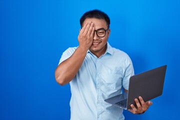 Chinese young man using computer laptop covering one eye with hand, confident smile on face and surprise emotion.