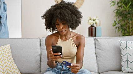 African american woman shopping with smartphone and credit card sitting on sofa at home
