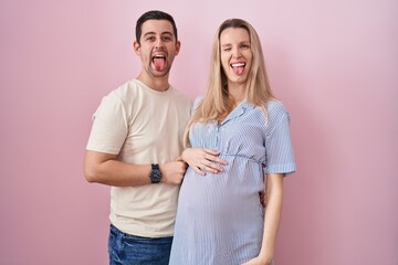 Young couple expecting a baby standing over pink background sticking tongue out happy with funny...