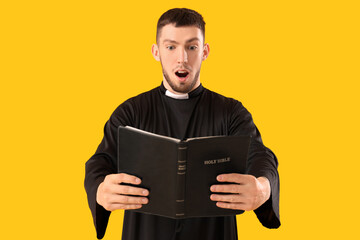 Shocked young priest with Holy Bible on yellow background