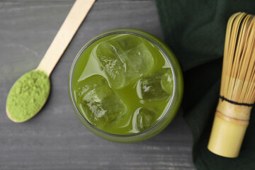 Glass of delicious iced green matcha tea, spoon with powder and bamboo whisk on grey wooden table,...