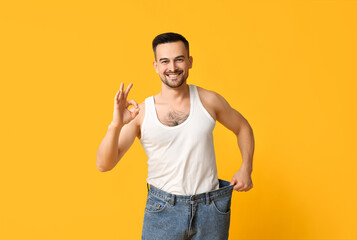 Handsome sporty happy man in loose jeans showing OK gesture on yellow background. Weight loss concept
