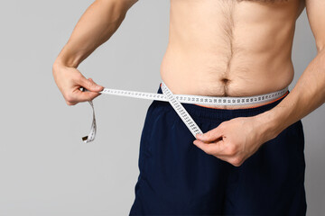 Handsome sporty young man measuring his waist on grey background, closeup. Weight loss concept