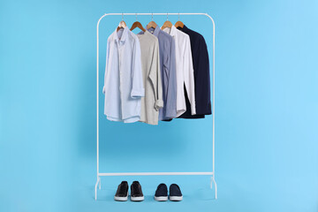 Rack with stylish clothes on wooden hangers and sneakers against light blue background