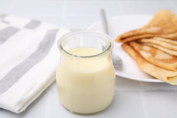 Tasty condensed milk in jar and crepes on white table, closeup