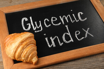 Small chalkboard with words Glycemic Index and croissant on wooden table, closeup