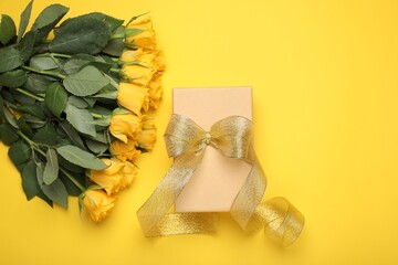Gift box with golden bow and bouquet of beautiful roses on yellow background, flat lay. Space for text