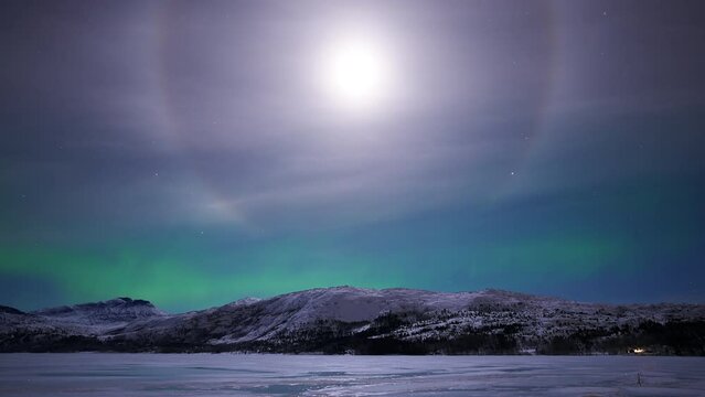 Moon surrounded by a halo and a distant aurora dancing above mountains on a cold night in early December.