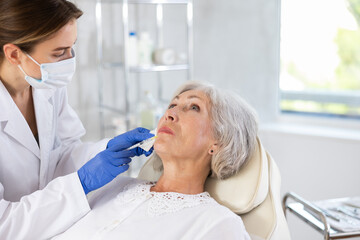 Fototapeta premium Serious old lady with plastic surgery in studio for skincare, cosmetic and wrinkles filler. The expert beautician is injecting.
