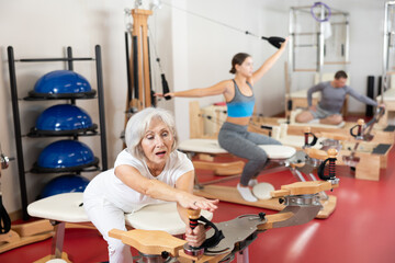 Elderly woman in sportswear stretches her arms and shoulders using pilates machine..