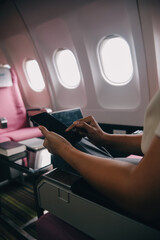 Asian woman passenger sitting in airplane near window and reading news from social networks or using travel applications in smartphone