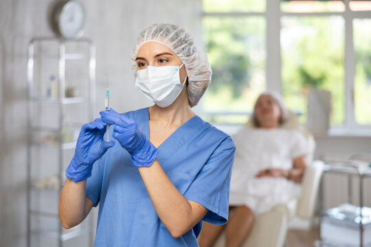 Young female doctor wearing medical uniform, mask and gloves preparing injection for female patient at treatment room