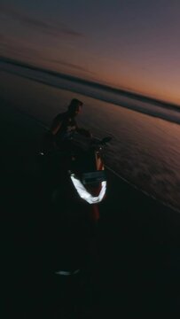 Vertical video. Aerial view man motorcyclist riding red bike hand shaka night sunset beach sea tropical landscape. FPV sport drone shot evening dusk coast flying over endless tide calm foam wave ocean