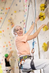 Tuinposter Determined senior woman doing her best at climbing artificial wall © JackF